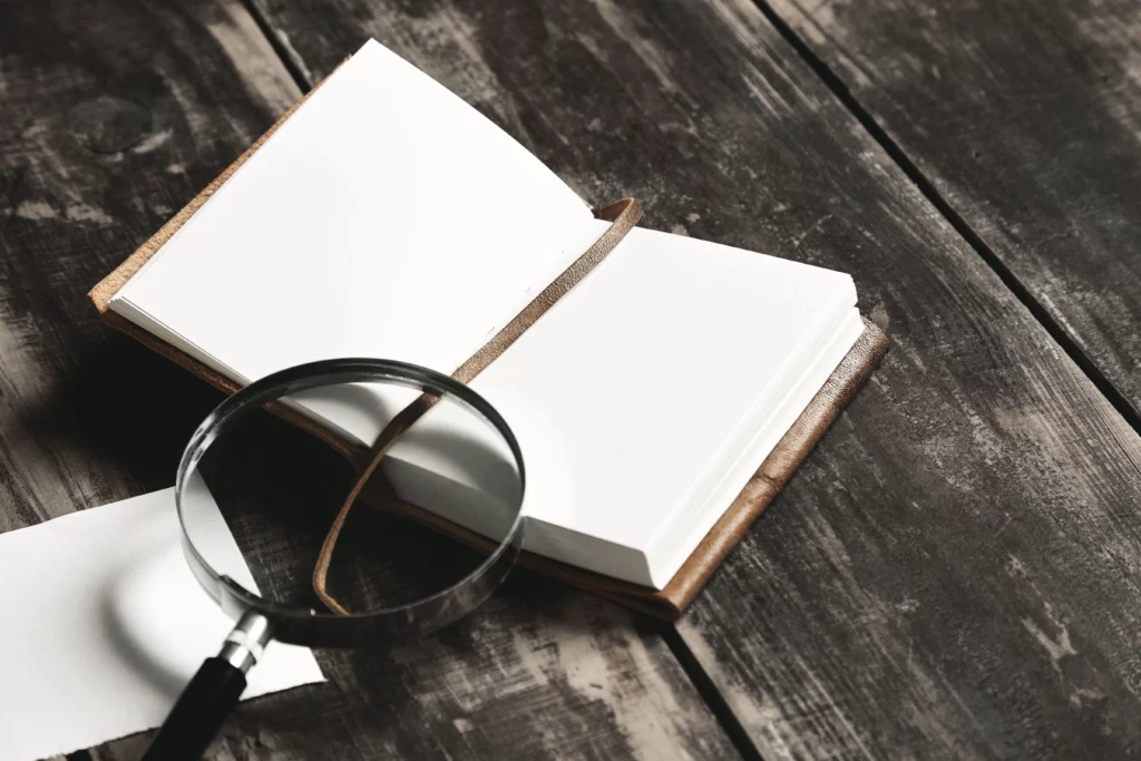 C'est l'en-tête de la page Contact. Il y a un carnet en cuir marron ouvert sur un table en bois façon rustique. Une loupe est également posée sur le carnet